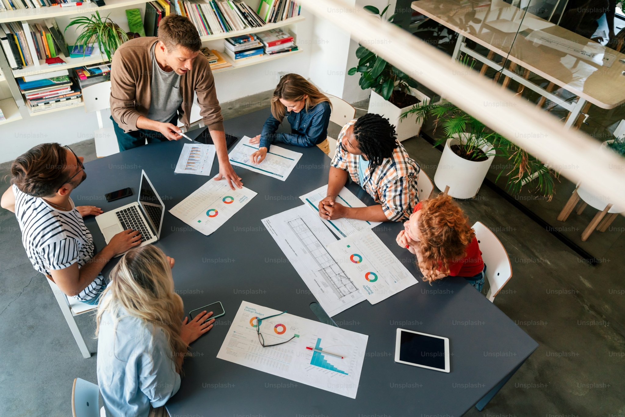 A group of colleague collaborate over a co-working table adorned with papers displaying data, a laptop and tablet device.
